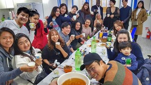 Un grupo de personas sentadas alrededor de una mesa con comida y bebidas, en una habitación con paredes blancas y una puerta al fondo.