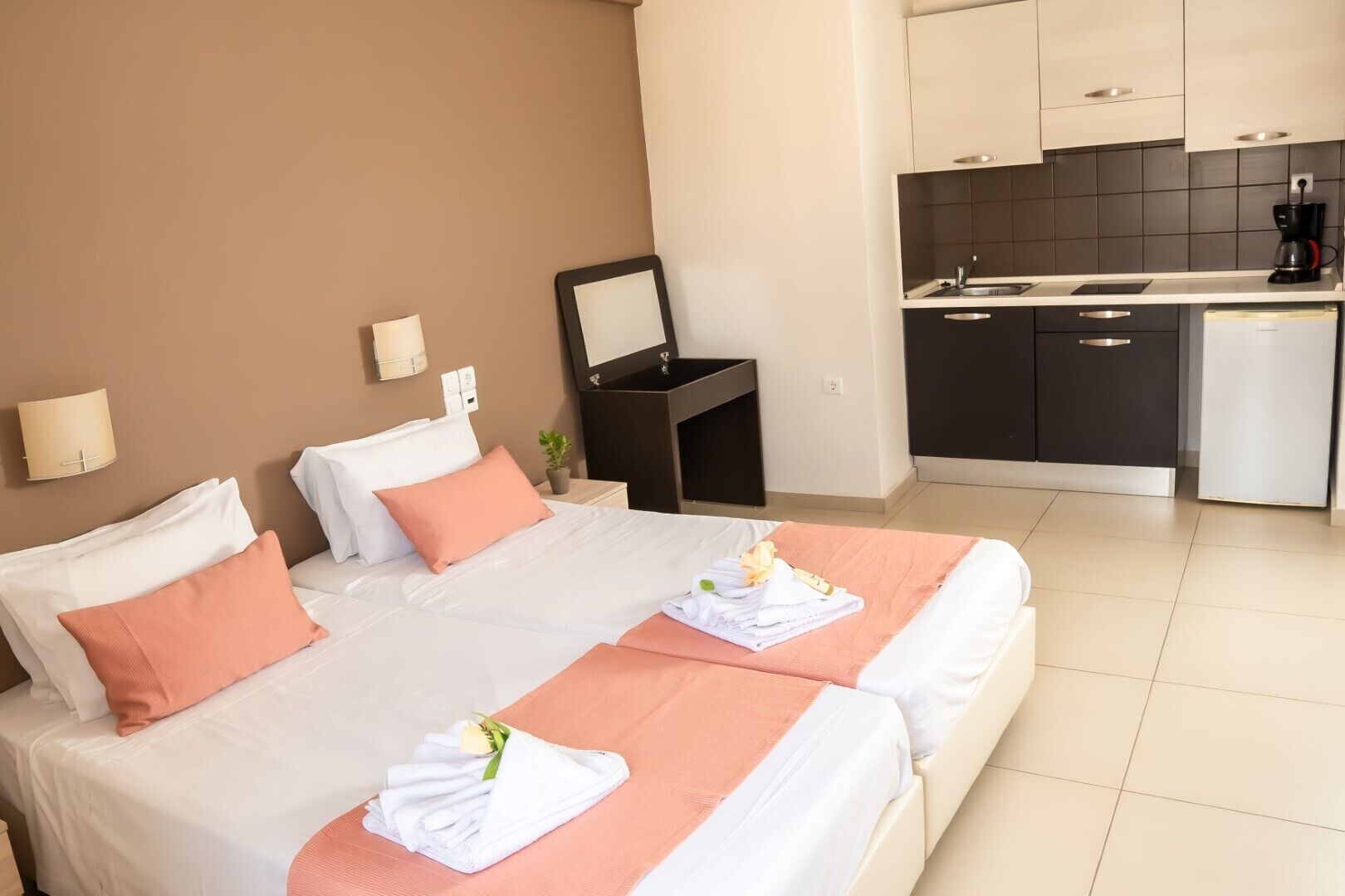A hotel room with a bed, a bedside table, a mirror, a small potted plant, and a kitchenette.