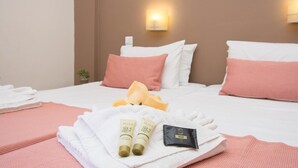 Una cama bien hecha con almohadas blancas y rosadas, una toalla blanca doblada y artículos de tocador, incluyendo dos tubos de jabón de aceite de oliva y una pequeña pastilla de jabón.