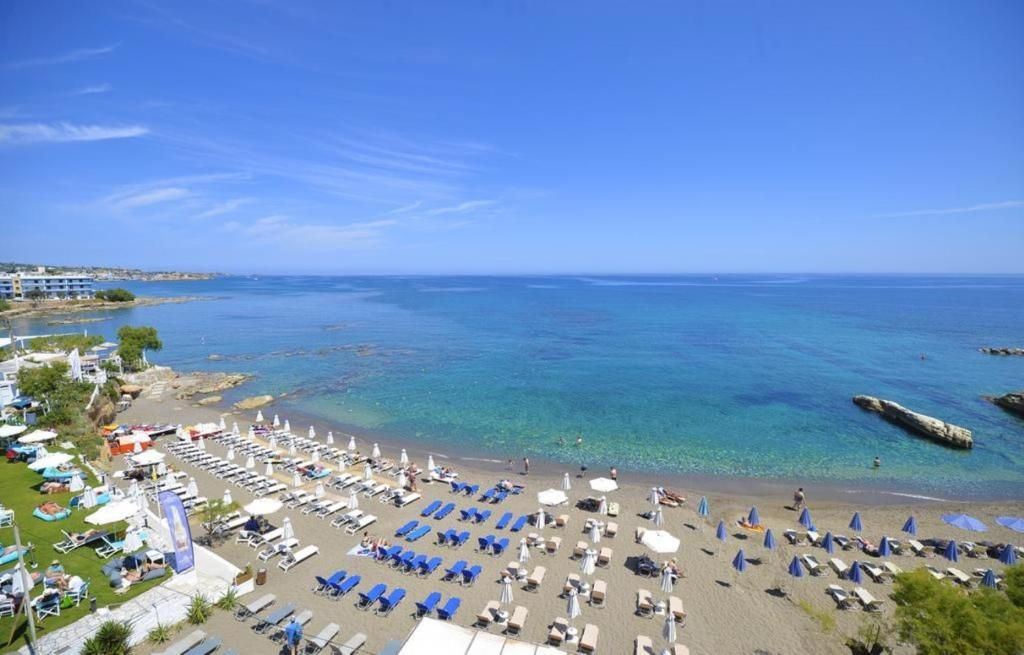 Una playa con tumbonas y sombrillas, un mar azul cristalino y edificios al fondo.