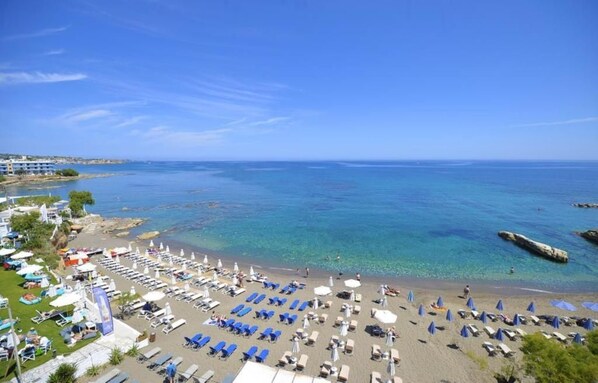 Uma praia com espreguiçadeiras e guarda-sóis, mar azul cristalino e edifícios ao fundo.