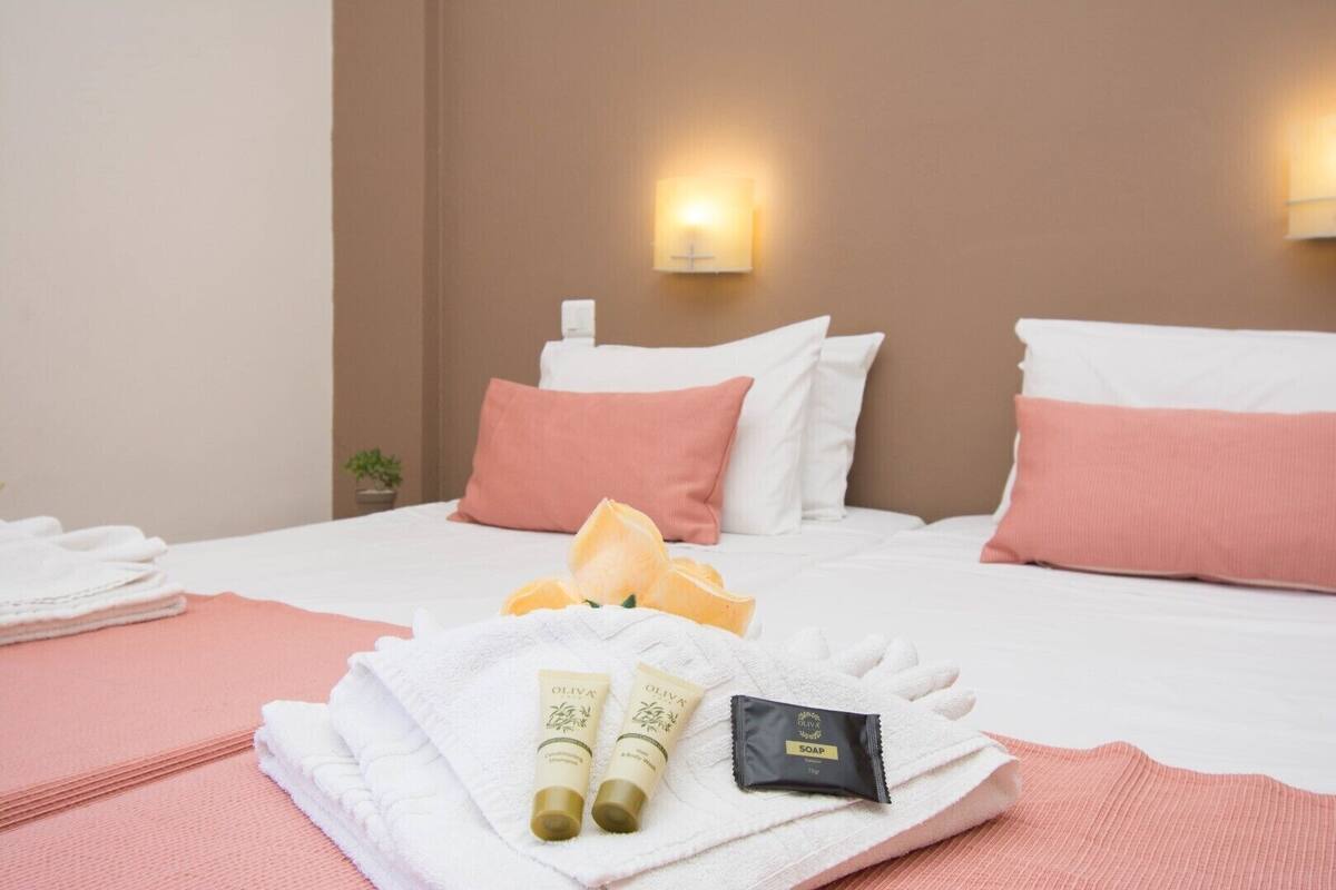 Ein Hotelzimmer mit zwei Betten, jeweils mit weißer und rosa Bettwäsche, einem ordentlich gefalteten weißen Handtuch und Pflegeprodukten, darunter zwei Flaschen und eine Seife.