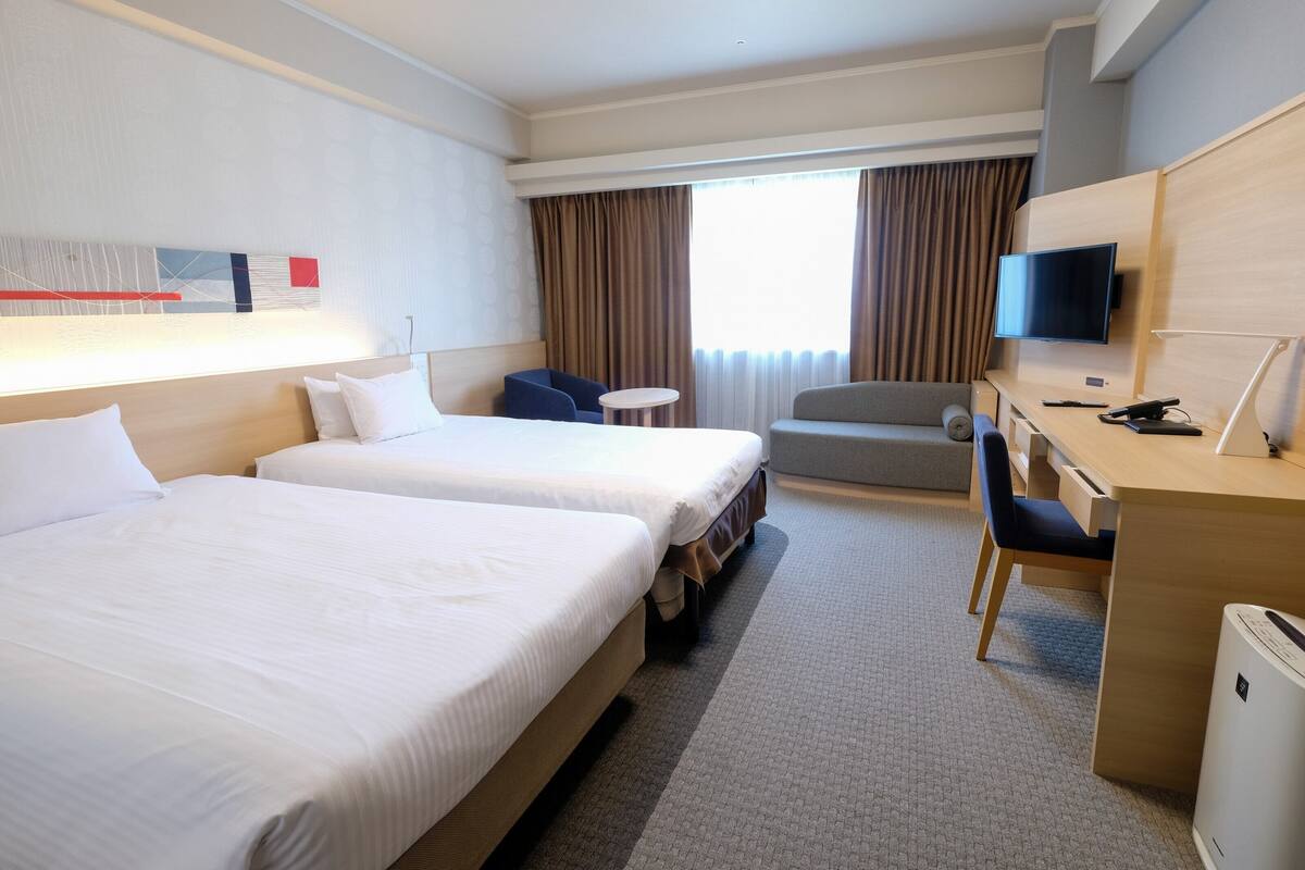 A hotel room with two beds, a desk, a chair, a television, and a window with curtains.
