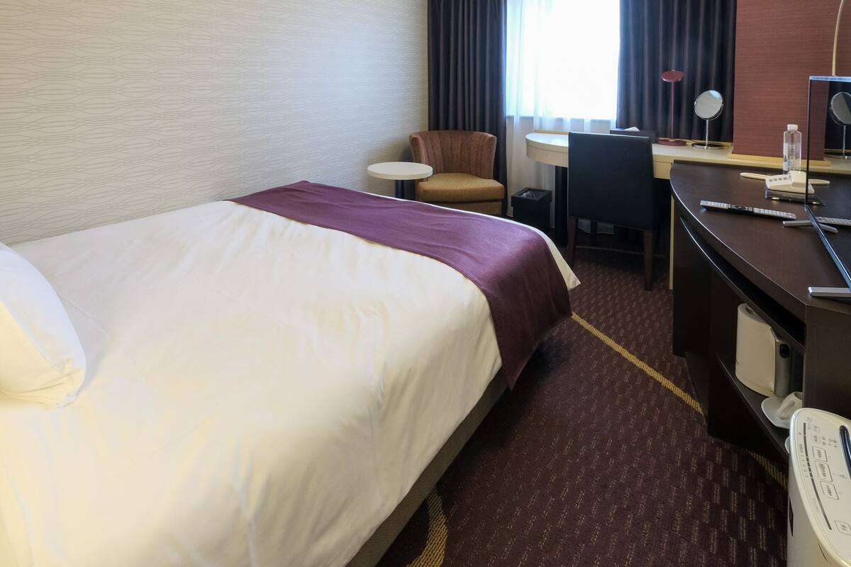 A hotel room with a large bed, a desk with a chair, a small table, and a window with curtains.