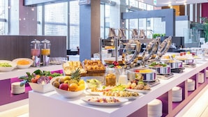 A buffet with various food items and beverages, including fruits, salads, and pastries, in a modern hotel setting with large windows and a bright interior.