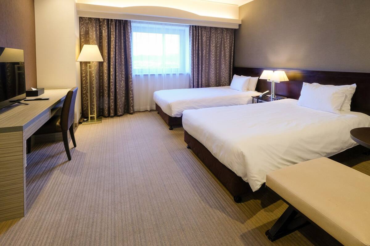 A hotel room with two beds, a desk, a chair, a lamp, and a window with curtains.