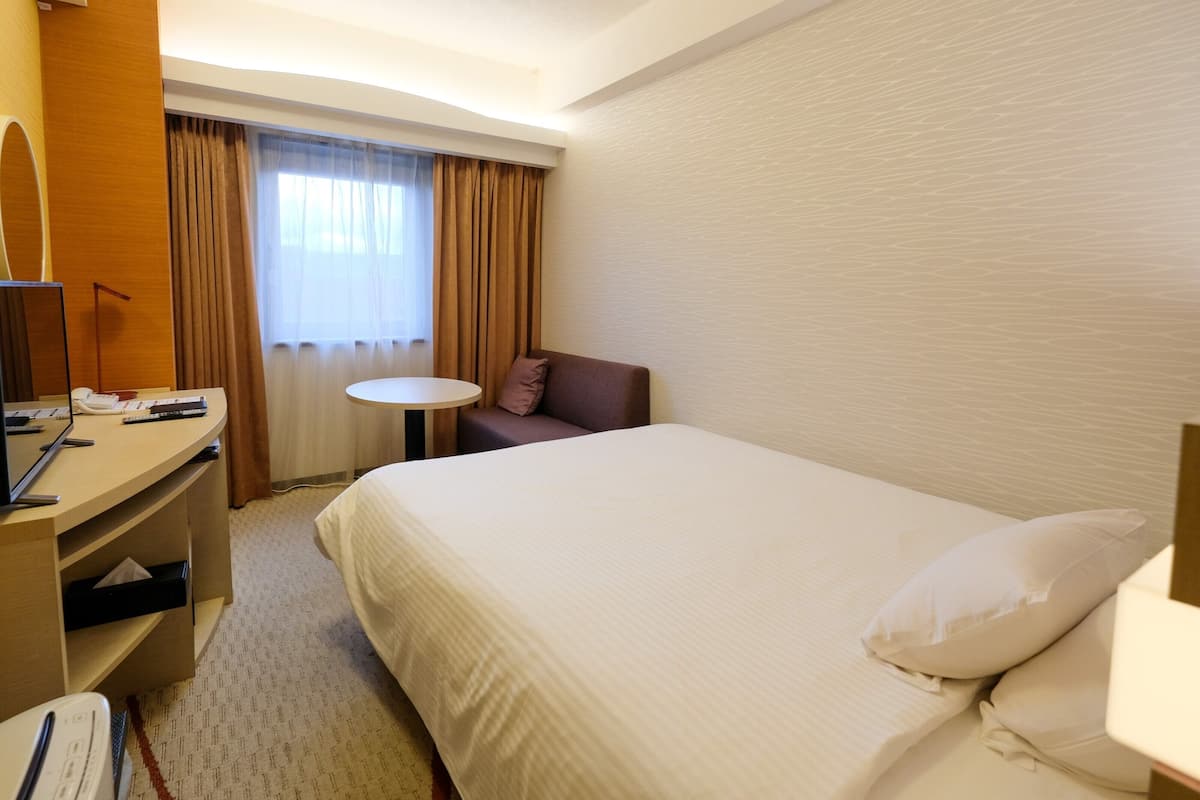A hotel room with a bed, a desk with a computer, a chair, a small table, and a window with curtains.
