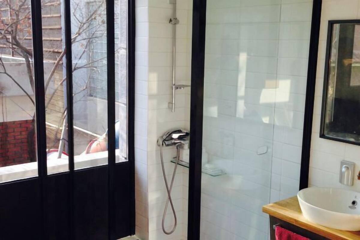A bathroom with a glass shower enclosure, a white sink, and a window with a view of a brick building outside.