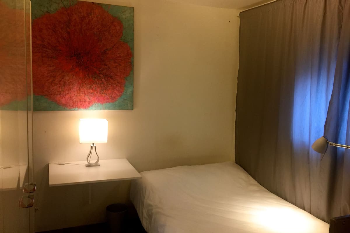 A small, single-bed room with a white bedside table, a lamp, a large floral artwork on the wall, and a window with curtains.