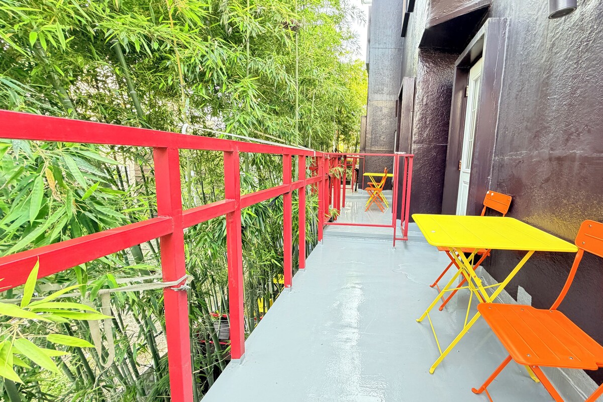 A narrow outdoor corridor with a red railing, a yellow table, and orange chairs.
