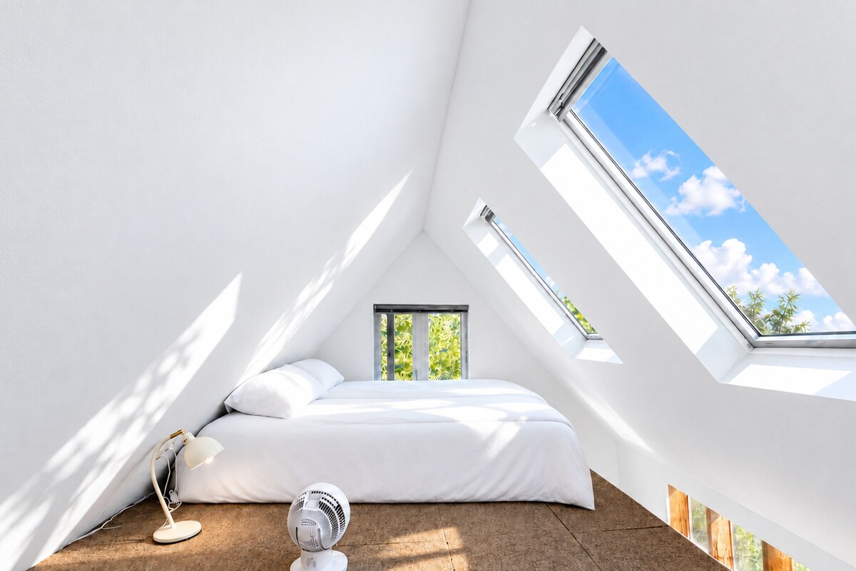 A bedroom with a sloped ceiling, a bed, a desk, a chair, and a window with a view of greenery.
