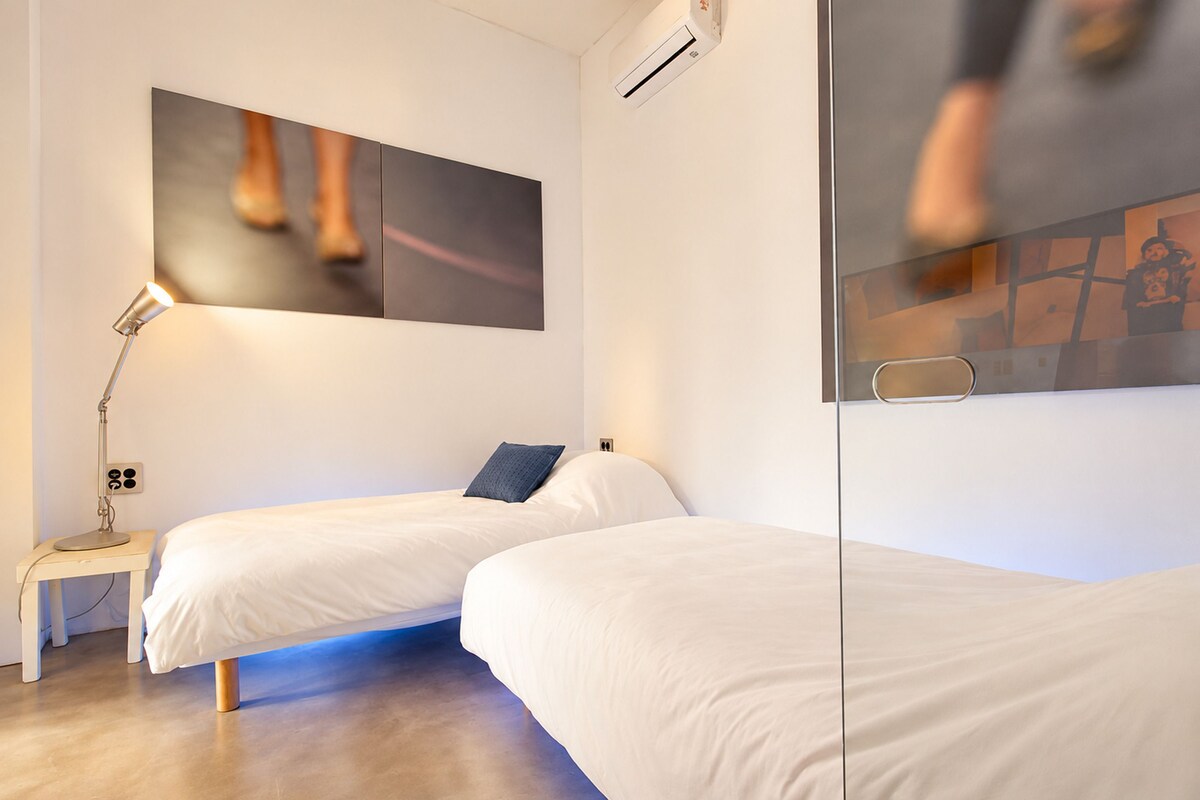 A hotel room with two single beds, a bedside table with a lamp, a wall-mounted air conditioning unit, and a large window.