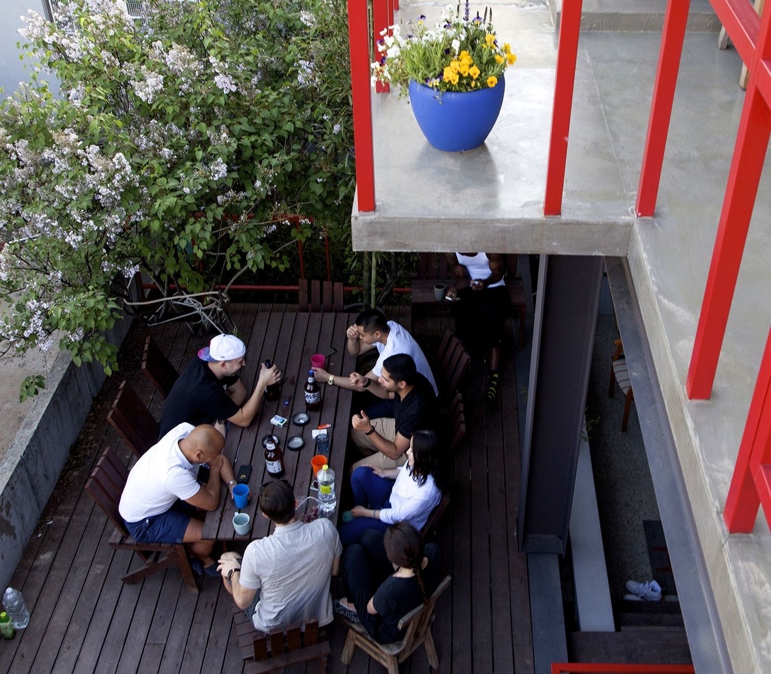 A group of people seated at a wooden table outdoors, with a large planter with flowers above them.