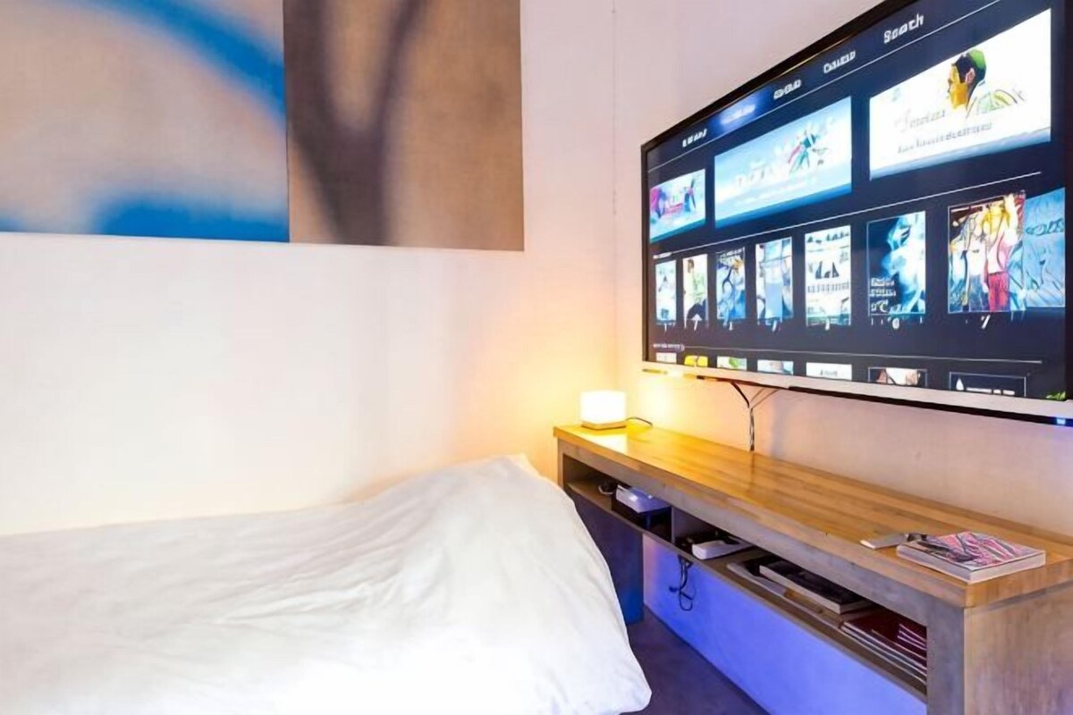 A hotel room with a bed, a bedside table with a lamp, and a wall-mounted TV displaying various channels.
