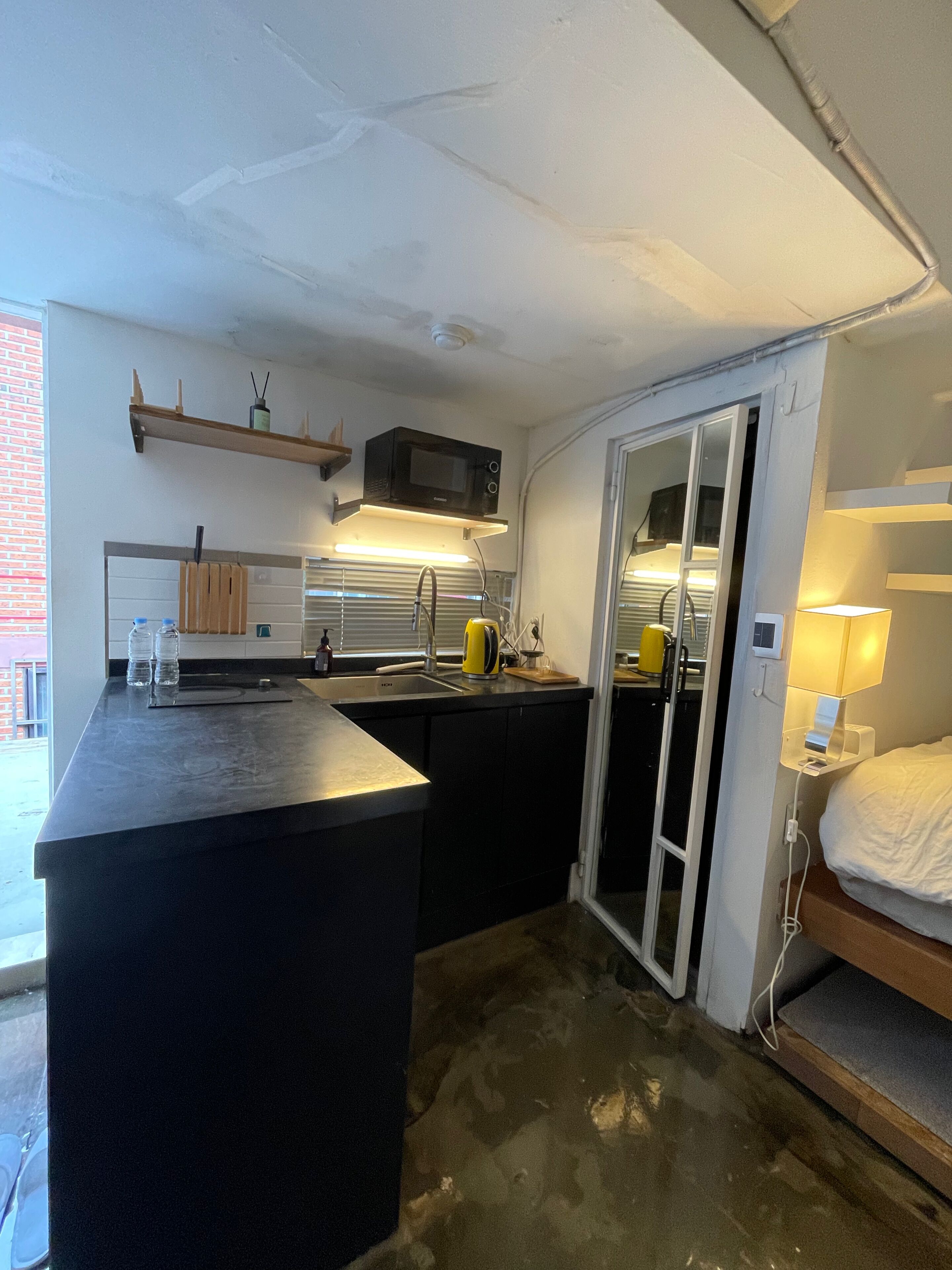 A compact kitchen with a black countertop, a microwave, and a sink. There is a small wooden shelf above the microwave and a window with blinds.
