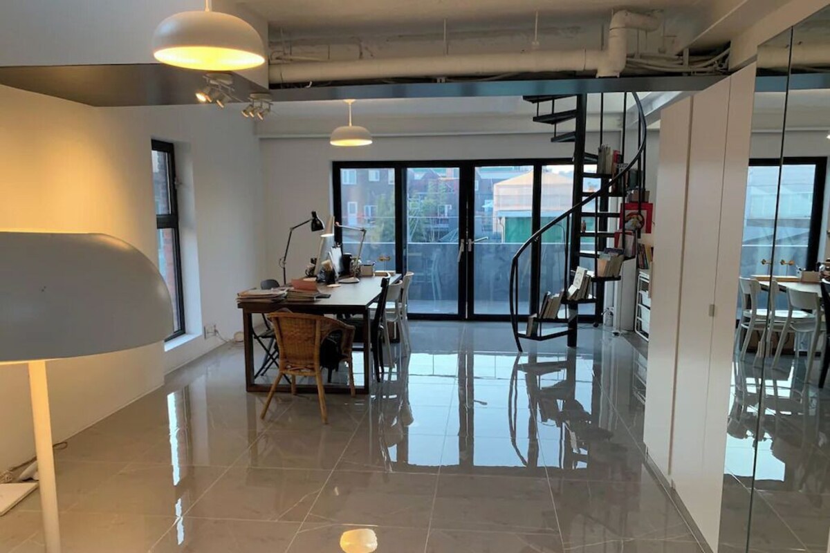 A spacious office with a polished floor, a desk with chairs, a staircase, and large windows offering a view of the outside.