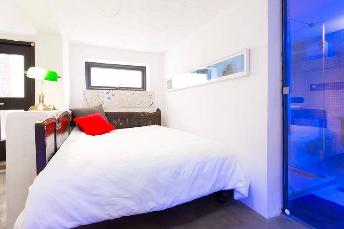 A bedroom with a bed, a window, a desk with a lamp, and a door leading to a balcony.