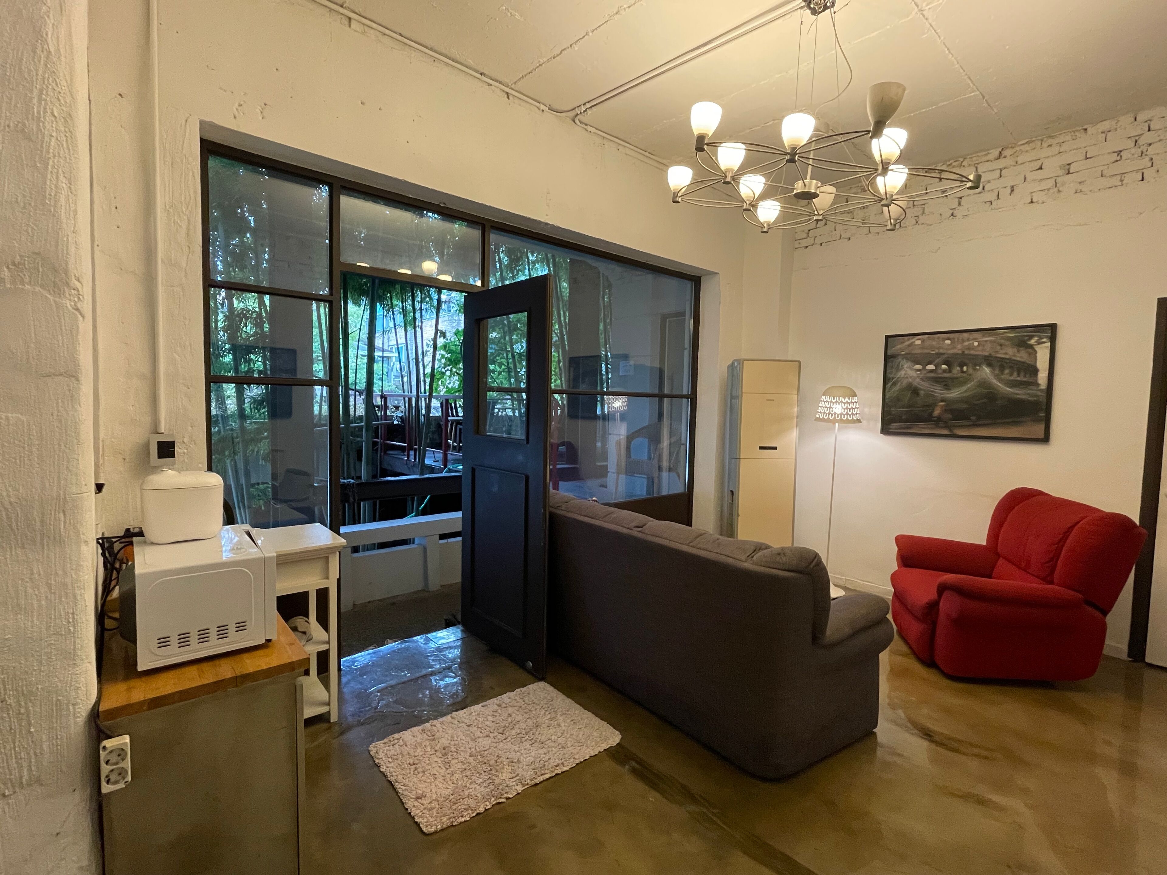 Una habitación moderna con un ventanal grande, un sofá seccional gris, un sillón rojo, un escritorio blanco con un ordenador y una araña.