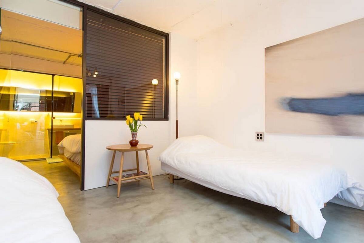A modern hotel room with two single beds, a small round table with a vase of flowers, and a large window with blinds.