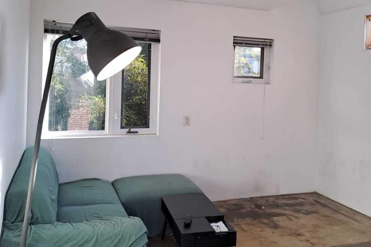 A room with a green sofa, a black desk, and a floor lamp. There are two windows, one with a view of greenery and another with a view of a building.