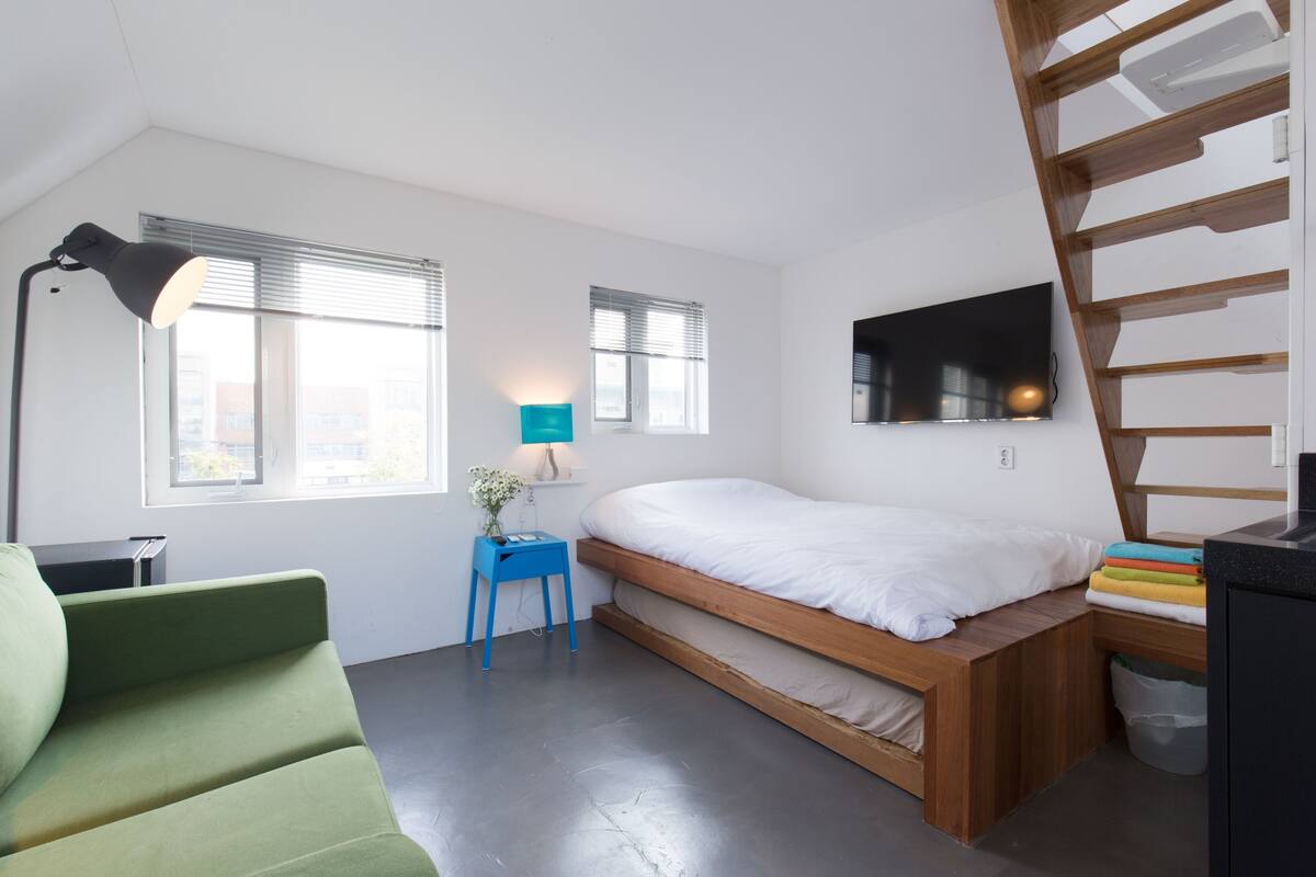 A modern bedroom with a loft bed, a desk with a lamp, a green chair, and a TV on the wall.