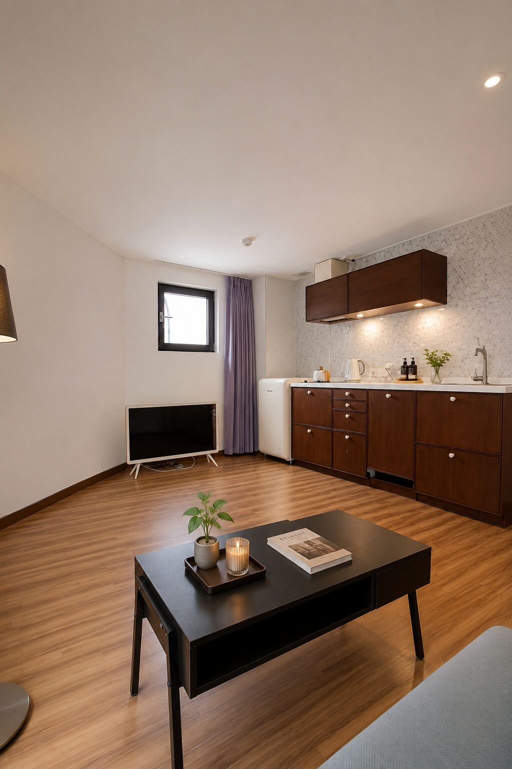 Una sala de estar moderna con un televisor de pantalla plana, una mesa de centro con una planta y una vela, un sofá y una cocina con armarios de madera y una ventana.