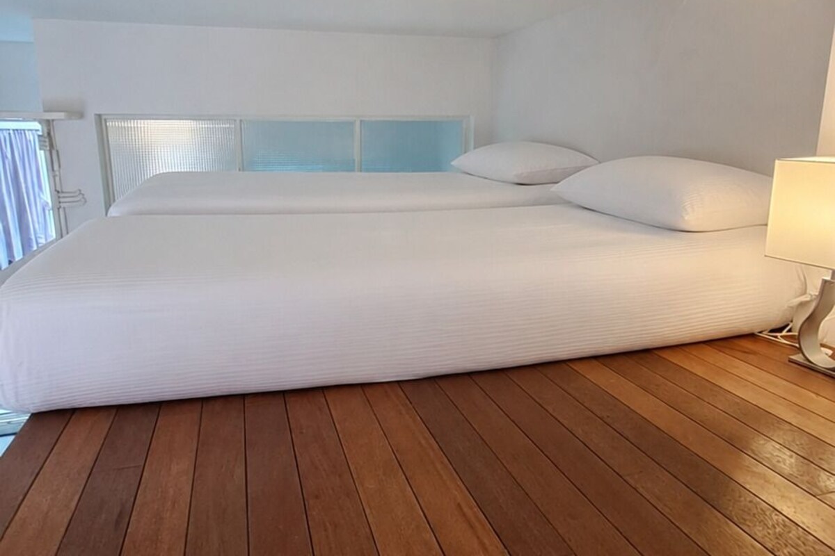 A room with a large bed, wooden flooring, a window with blinds, and a bedside table with a lamp.