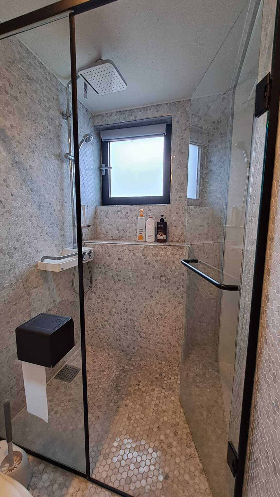Ducha con paredes de vidrio y ventana, un lavamanos pequeño y artículos de aseo en un estante.