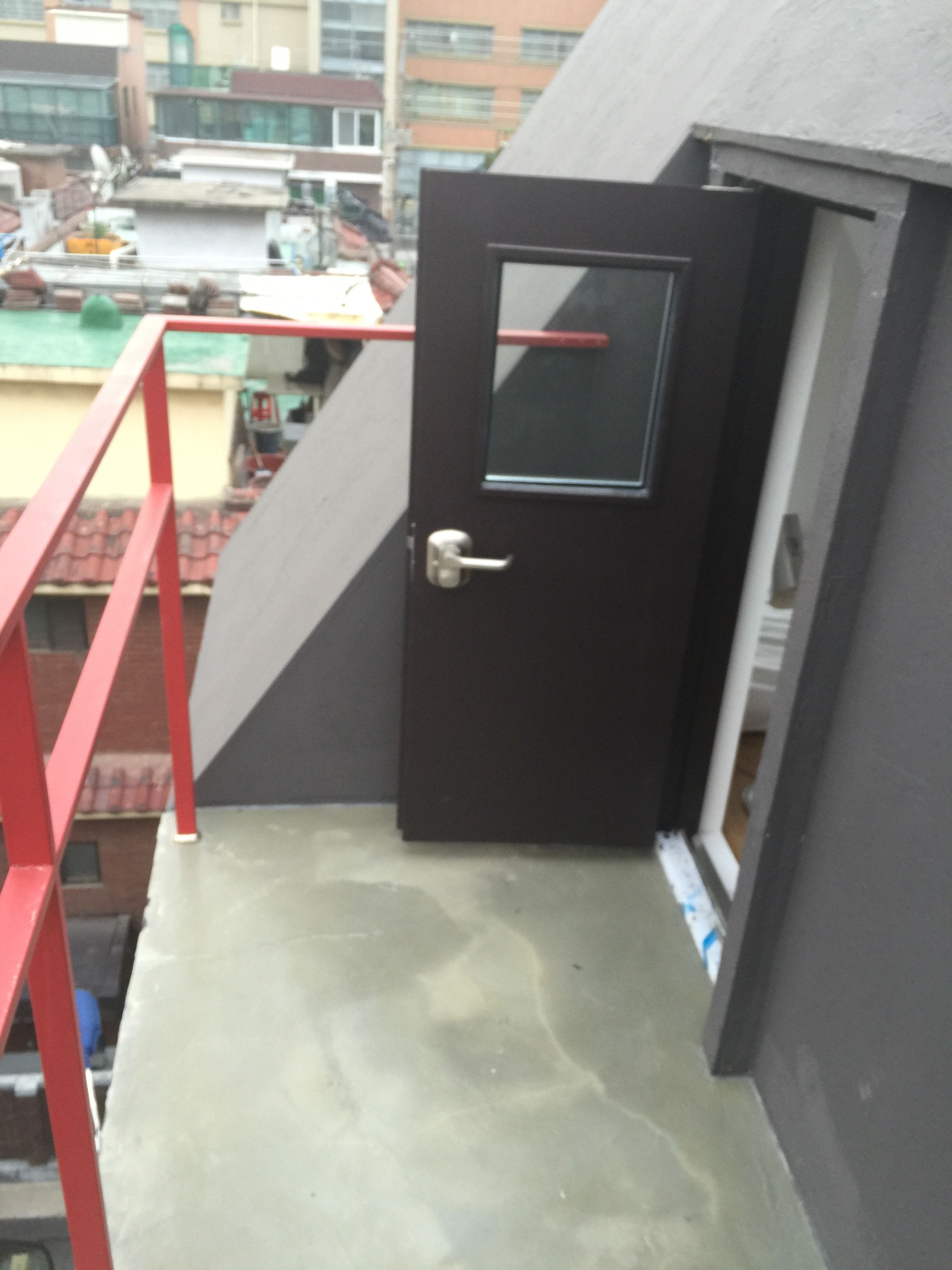 A black door with a small window, a red metal railing, and a concrete floor.