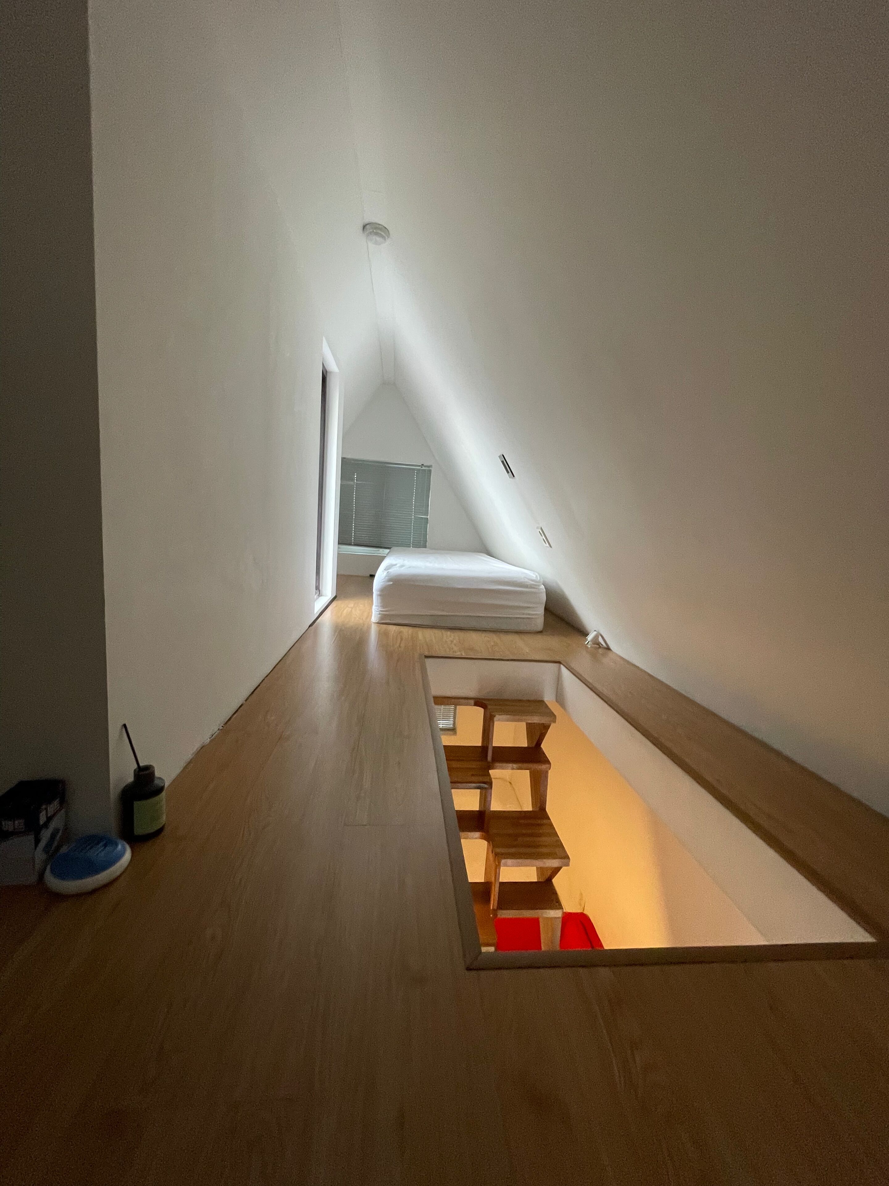 A narrow, sloped room with a wooden floor, a small window, and a bed with white bedding.