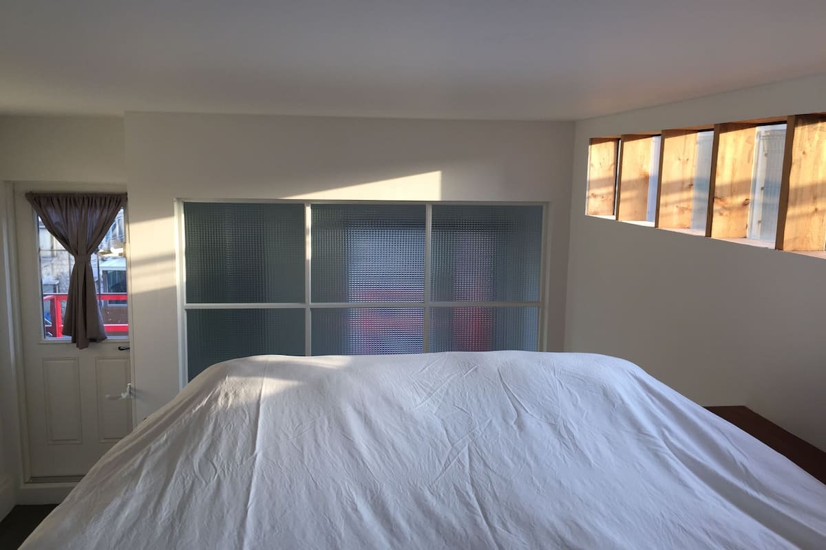 A bedroom with a bed, a window with blinds, a door, and a wall-mounted shelf.