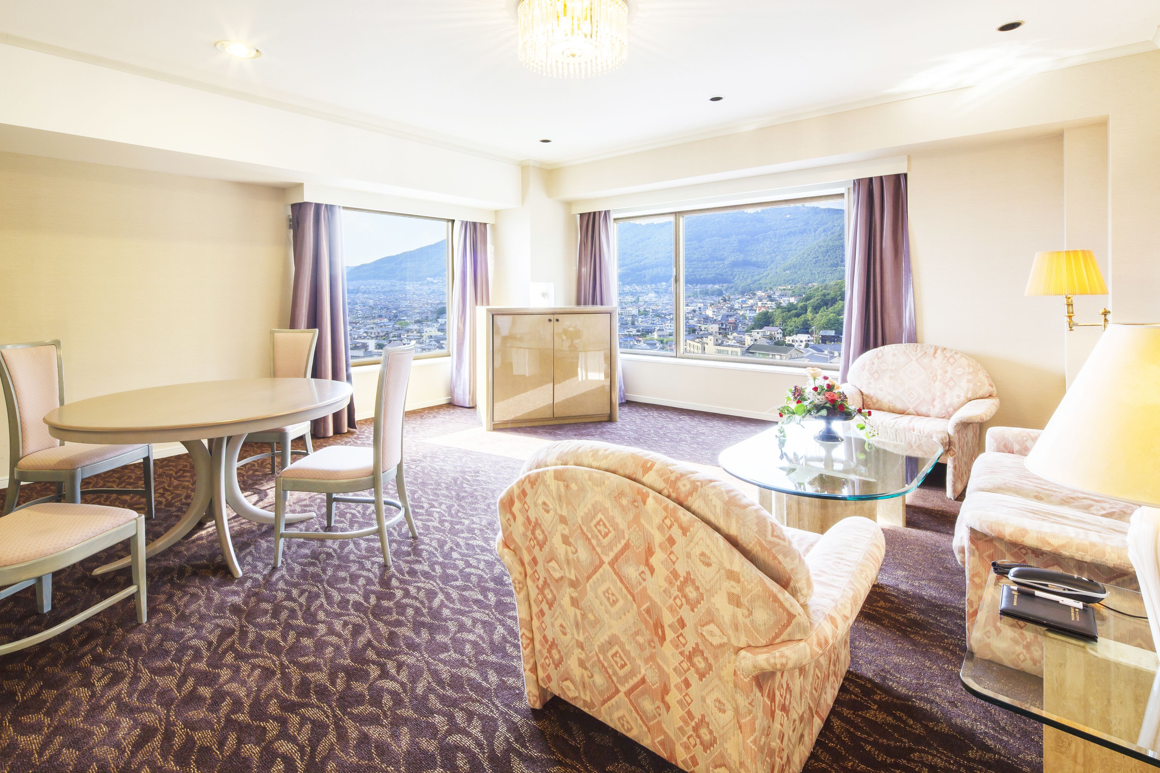Habitación de hotel con una mesa de comedor, dos sillones, un sofá, un televisor y vistas de un paisaje urbano a través de amplias ventanas.
