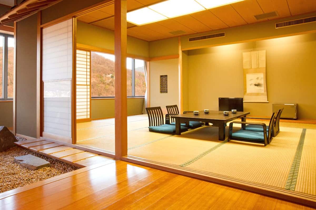 A traditional Japanese room with tatami flooring, a low table with cushions, and a view of a forested hillside.