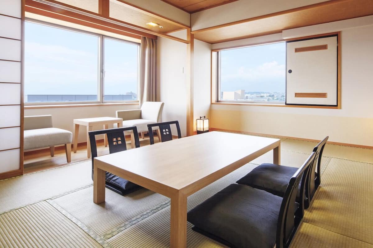 A room with a wooden table, black chairs, a sofa, tatami mats, and large windows offering a city view.