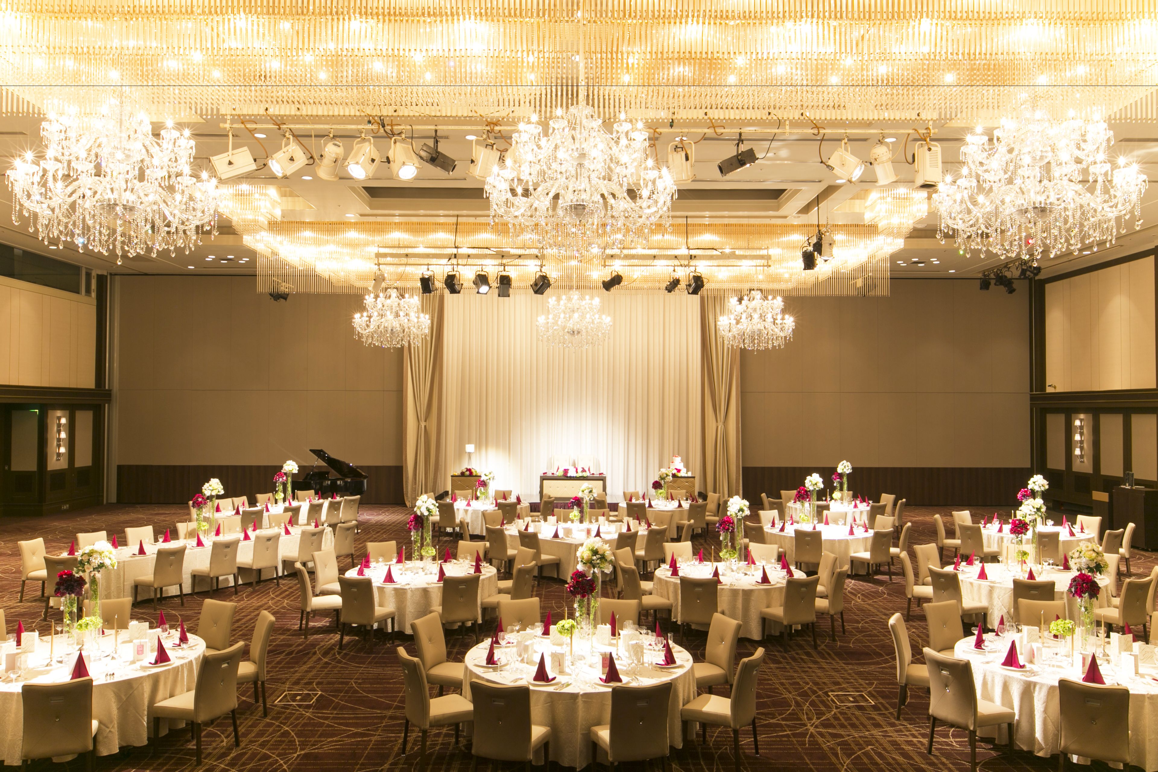 Un salón de banquetes con mesas redondas, sillas, candelabros y un escenario.