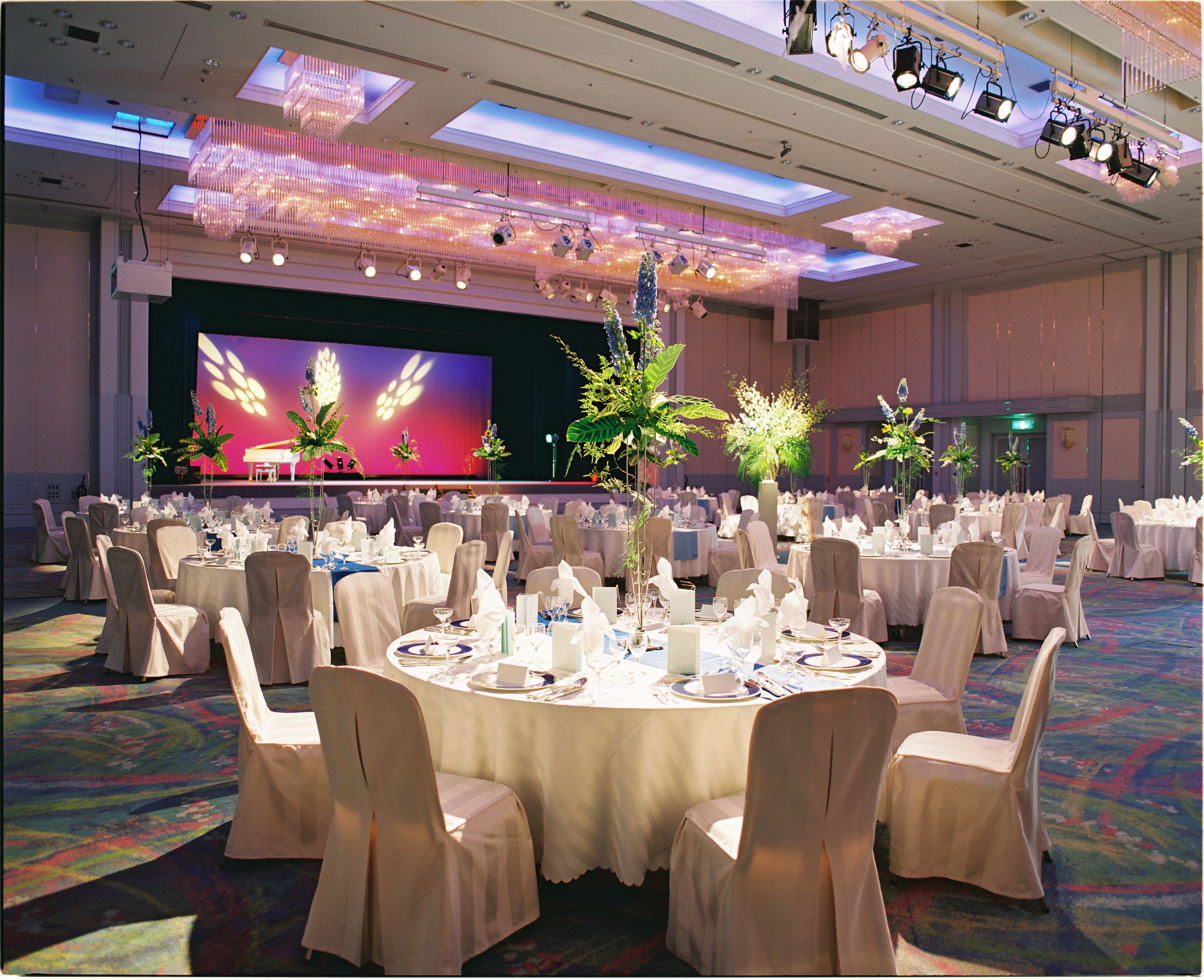 A banquet hall with round tables, chairs, and a stage with a large screen.