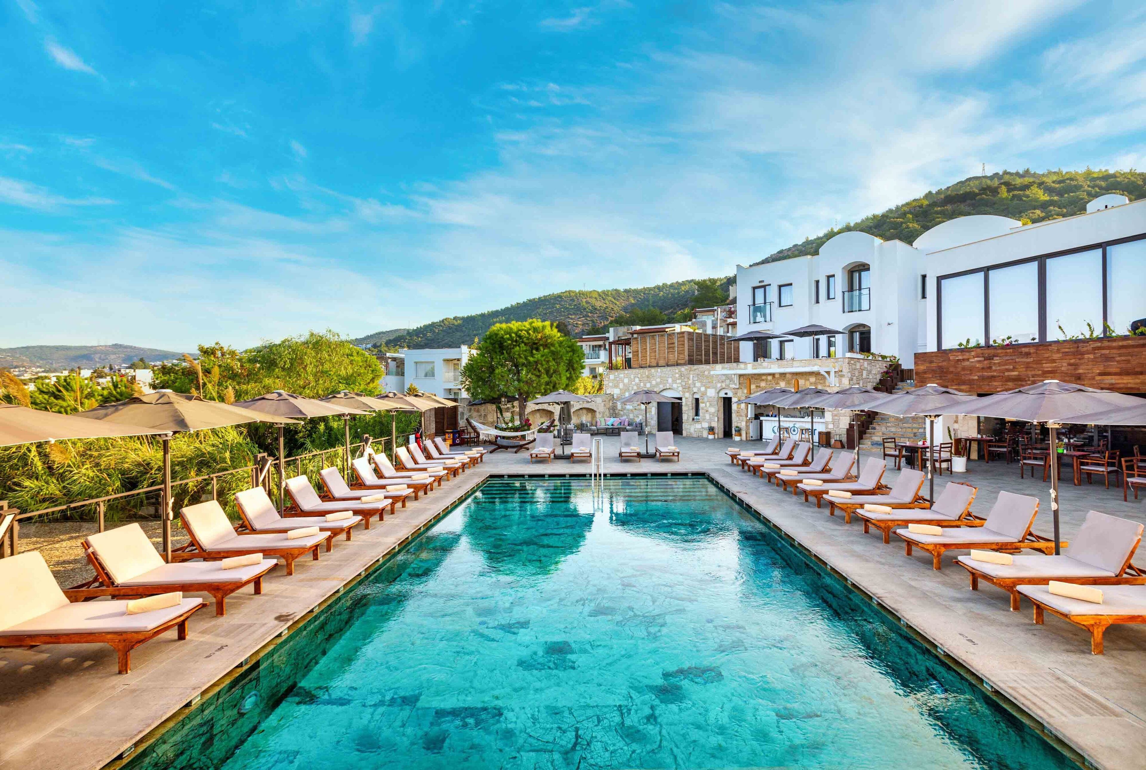 A poolside area with lounge chairs and umbrellas, a clear pool, and buildings in the background.