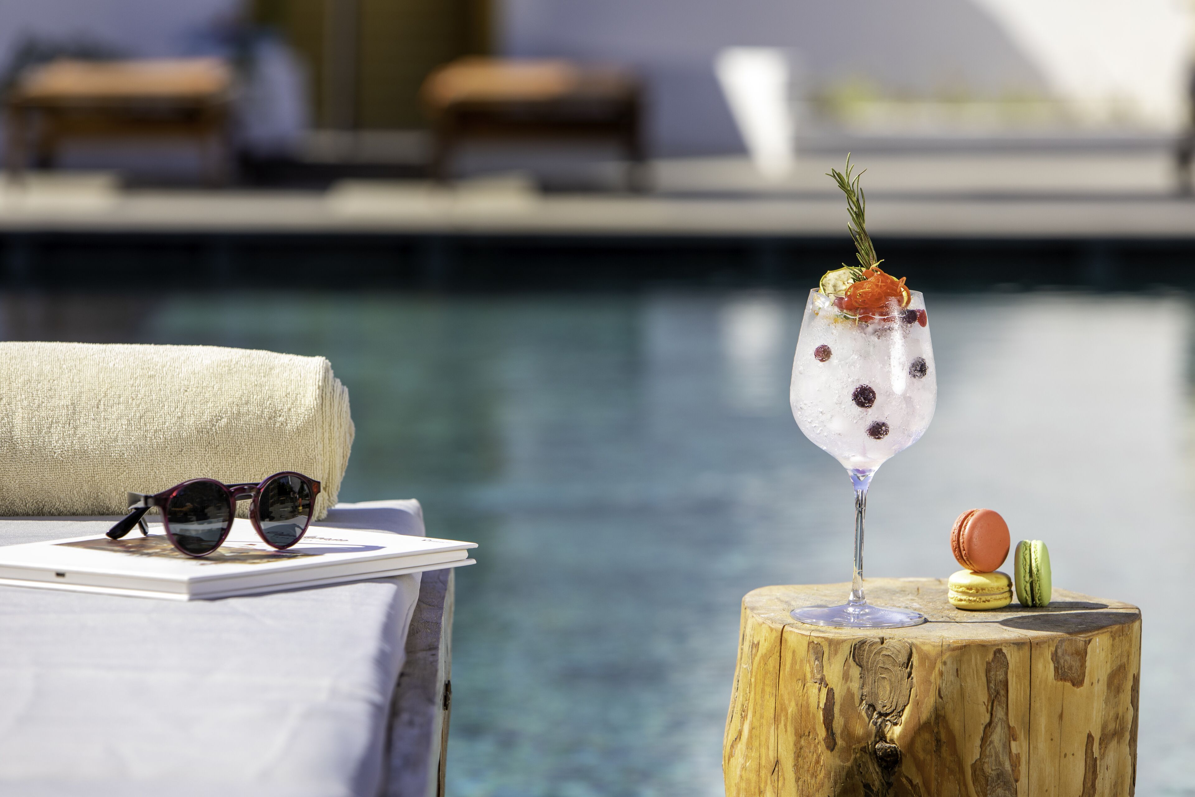 A glass of iced cocktail with a garnish on a wooden stump by a poolside.