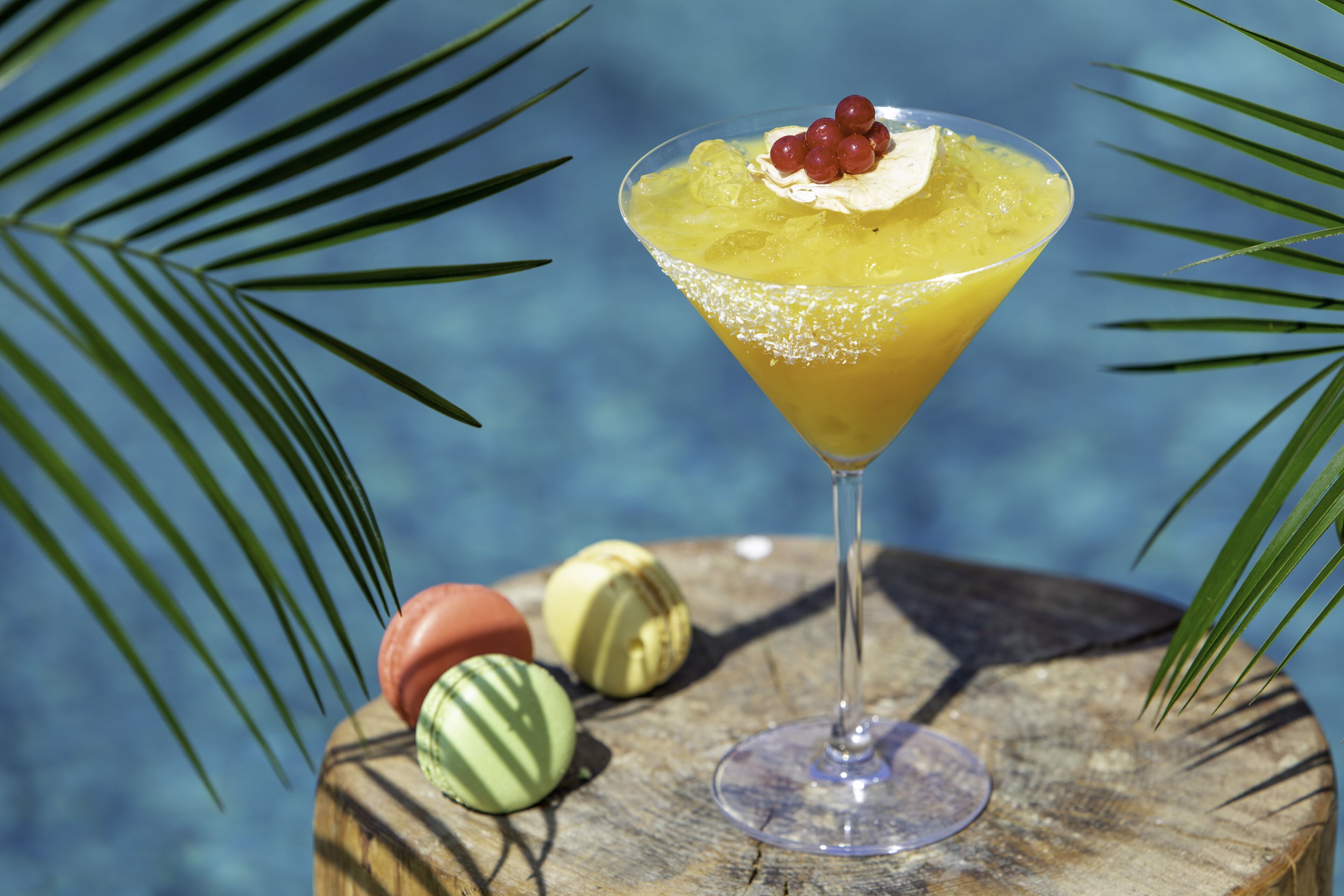 A cocktail in a martini glass with a garnish, placed on a wooden surface with palm leaves in the background.