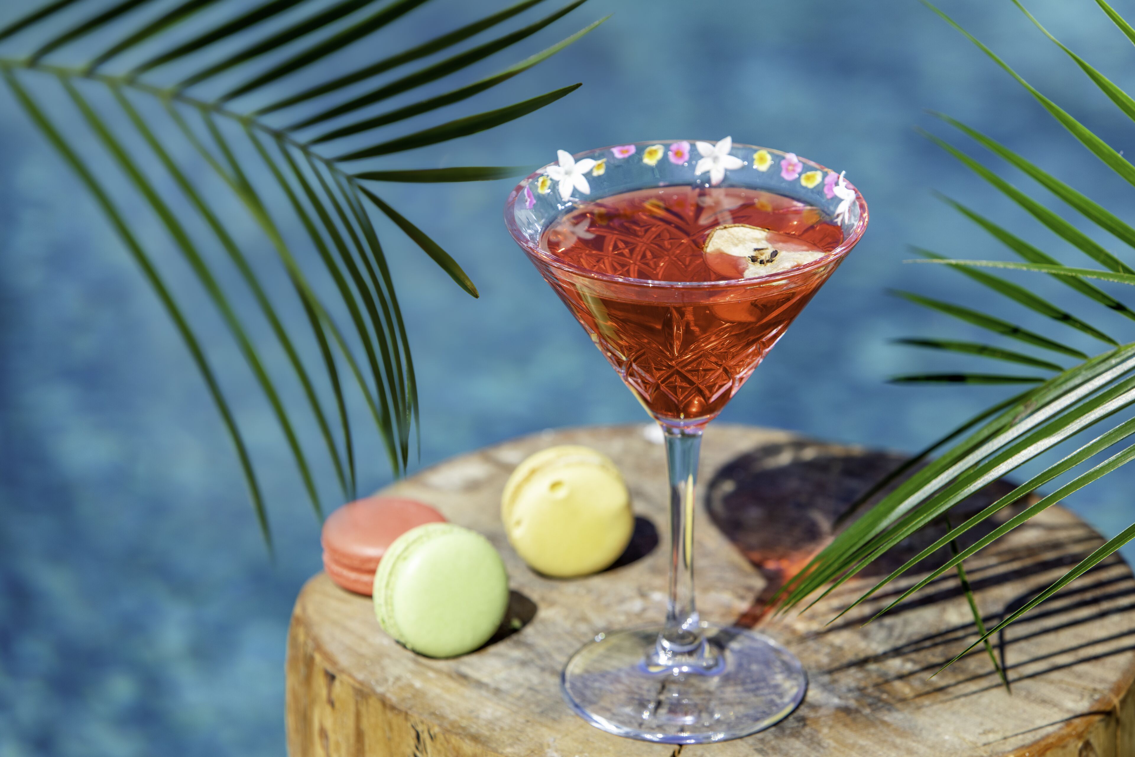 A cocktail in a glass with a floral rim, placed on a wooden surface with two colorful macarons beside it.