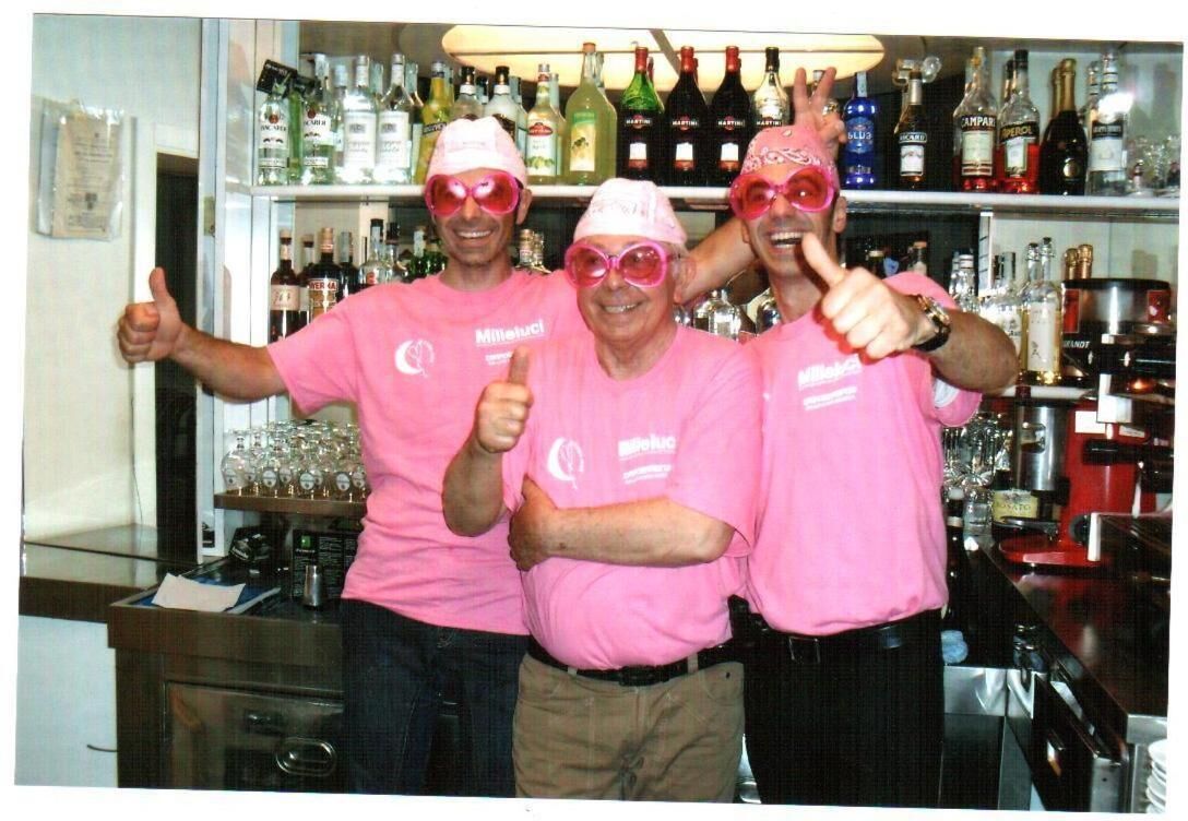 Trois personnes portant des chemises et des chapeaux roses lèvent le pouce dans un bar, avec des étagères remplies de bouteilles d’alcool.