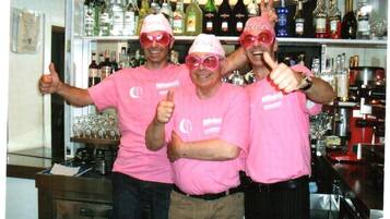Trois personnes portant des chemises et des chapeaux roses lèvent le pouce dans un bar, avec des étagères remplies de bouteilles d’alcool.