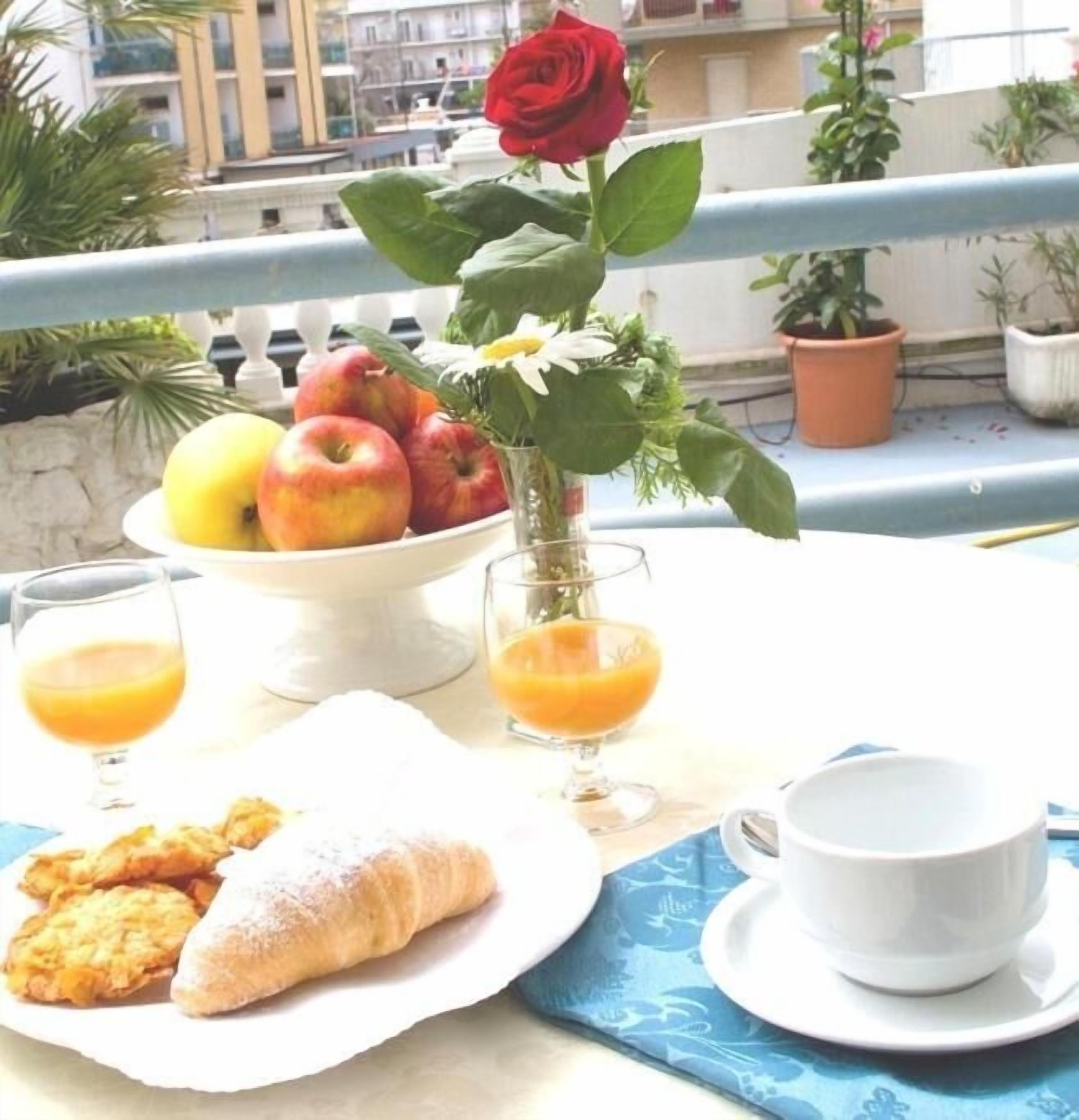 A table with a bowl of fruit, a rose, and a croissant on a balcony with potted plants and a railing.
