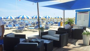 Een terras aan het strand met rieten meubels, blauw-witte parasols en een rij ligbedden op de achtergrond.
