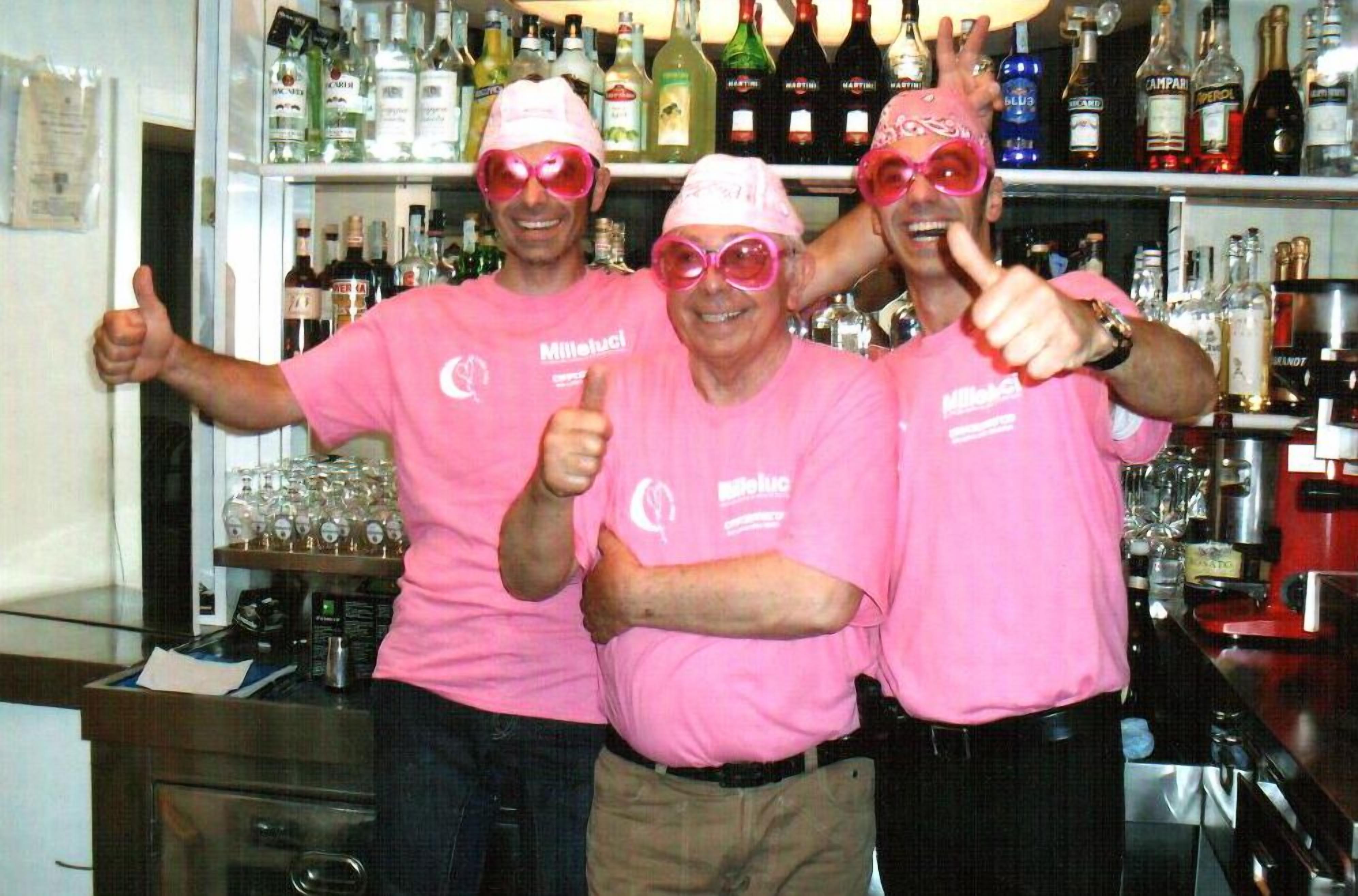 Three people in pink shirts with thumbs up, standing in a bar with shelves of liquor bottles behind them.