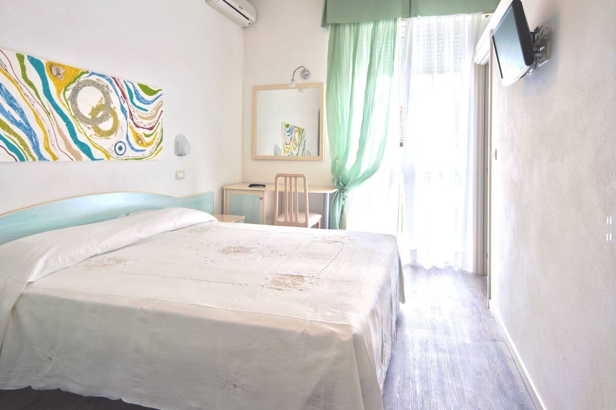 Une chambre d’hôtel avec un lit, un bureau, une chaise, un miroir et une fenêtre avec des rideaux.