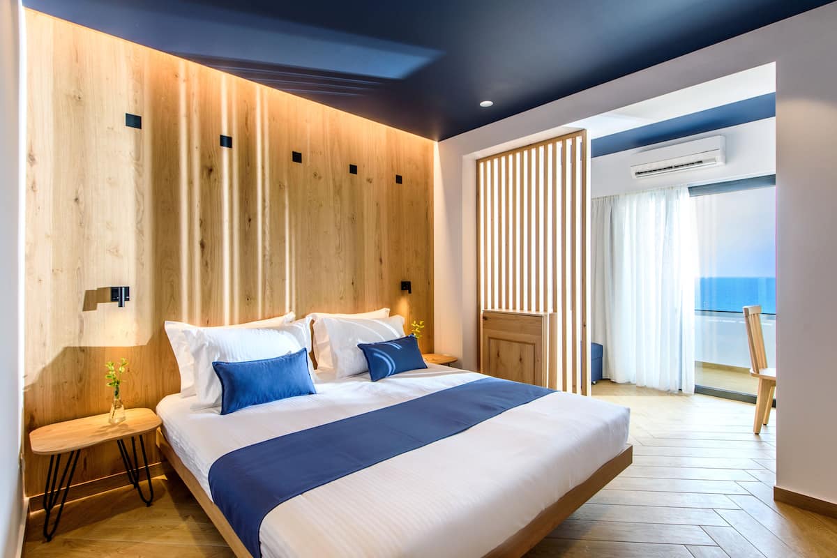 A modern hotel room with a large bed, wooden panel walls, a small table with a vase, and a view of the sea through a window.