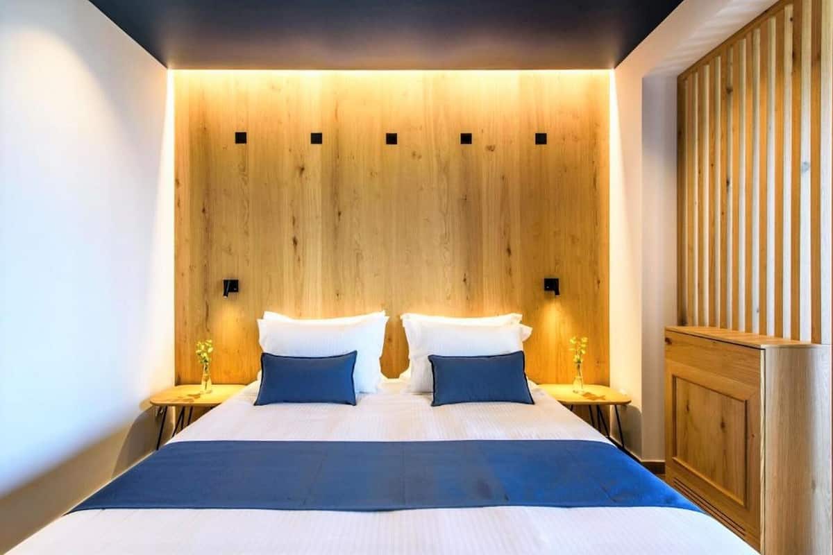 A modern hotel room with a large bed, wooden headboard, two bedside tables with lamps, and a wooden panel wall.
