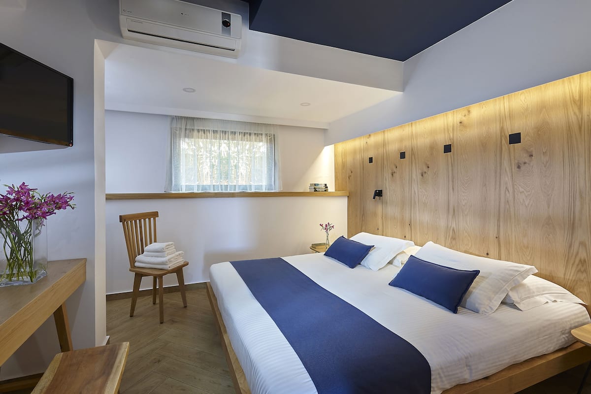 A modern hotel room with a large bed, wooden headboard, a desk with a chair, a window with sheer curtains, and an air conditioning unit on the ceiling.