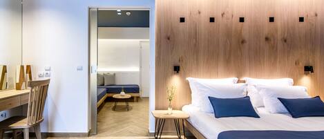 A modern hotel room with a wooden headboard, a large bed with blue bedding, a wooden desk, and a small side table with a vase.